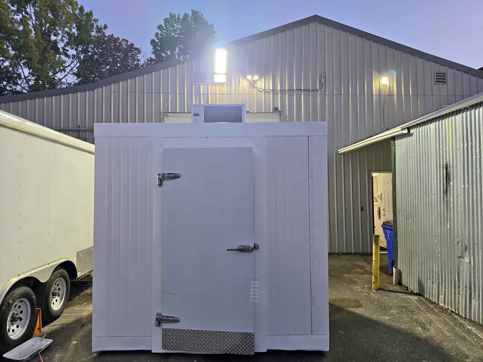 A white walk-in cooler with its refrigeration unit on top at dusk